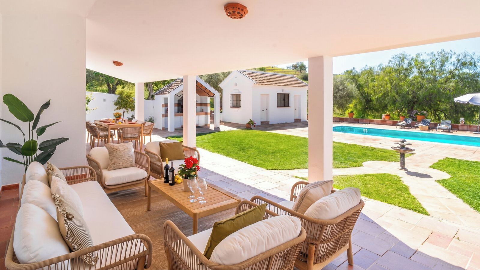 Ein überdachter Außenbereich mit Korbmöbeln, einem Tisch mit Wein und Blumen sowie Blick auf einen Swimmingpool und weiße Gebäude im Hintergrund.