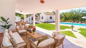 Ein überdachter Außenbereich mit Korbmöbeln, einem Tisch mit Wein und Blumen sowie Blick auf einen Swimmingpool und weiße Gebäude im Hintergrund.