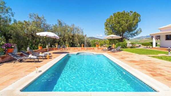 A rectangular swimming pool with a tiled edge, surrounded by lounge chairs and umbrellas, set in a landscaped garden with trees and a clear sky.