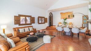 Ein geräumiges Wohnzimmer mit einer Sofagarnitur, einem Essbereich mit Stühlen, einem Kamin und einem großen Fenster mit Rollläden.
