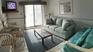 Ett vardagsrum med en ljusgrön soffa, ett soffbord och en turkos fåtölj. En tv är monterad på en hylla ovanför soffan, och det finns ett fönster med persienner till vänster.