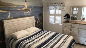Ett sovrum med en säng, en byrå, en spegel och en väggmålning som föreställer ett fyrtorn och hav.