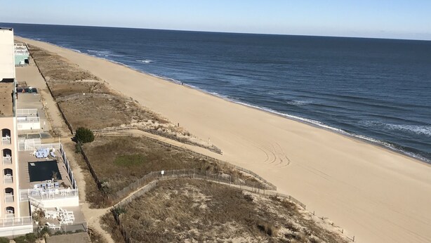 En strand med en sandstrand, en klarblå himmel och en byggnad med en balkong och ett utomhusområde med sittplatser.