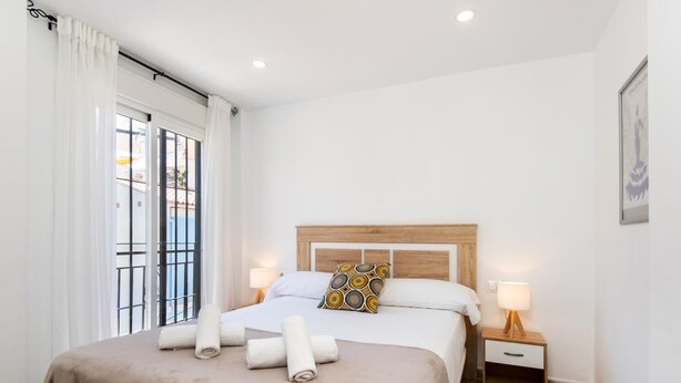 A neatly made bed with a brown cover, two bedside tables with lamps, a patterned pillow, and a window with curtains. - STREET ART FANTASY 1 APARTMENT (MALAGA)