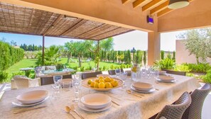 Uma área de refeições ao ar livre com mesa posta para quatro pessoas, cercada por cadeiras de vime, com vista para um jardim exuberante e um campo de golfe.