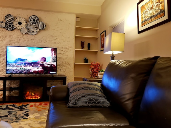 A living room with a leather sofa, a flat-screen TV, a fireplace, decorative wall art, and a bookshelf.
