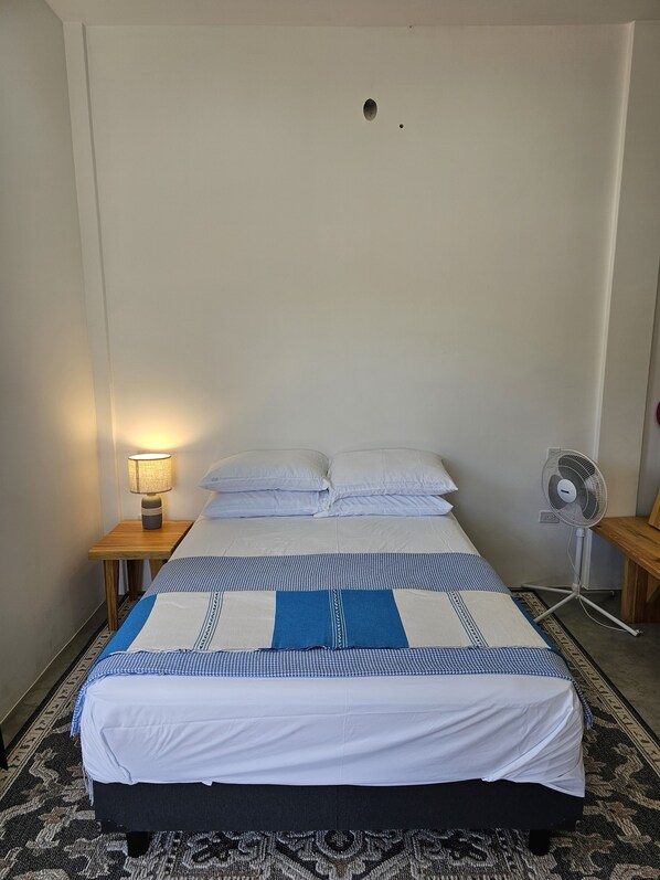 A single bed with a blue and white checkered bedspread, white pillows, a bedside table with a lamp, and a standing fan. - Private House with Ocean Views 10 mins to Cerritos Beach and Sand Pedrito Break! (Los Cerritos)