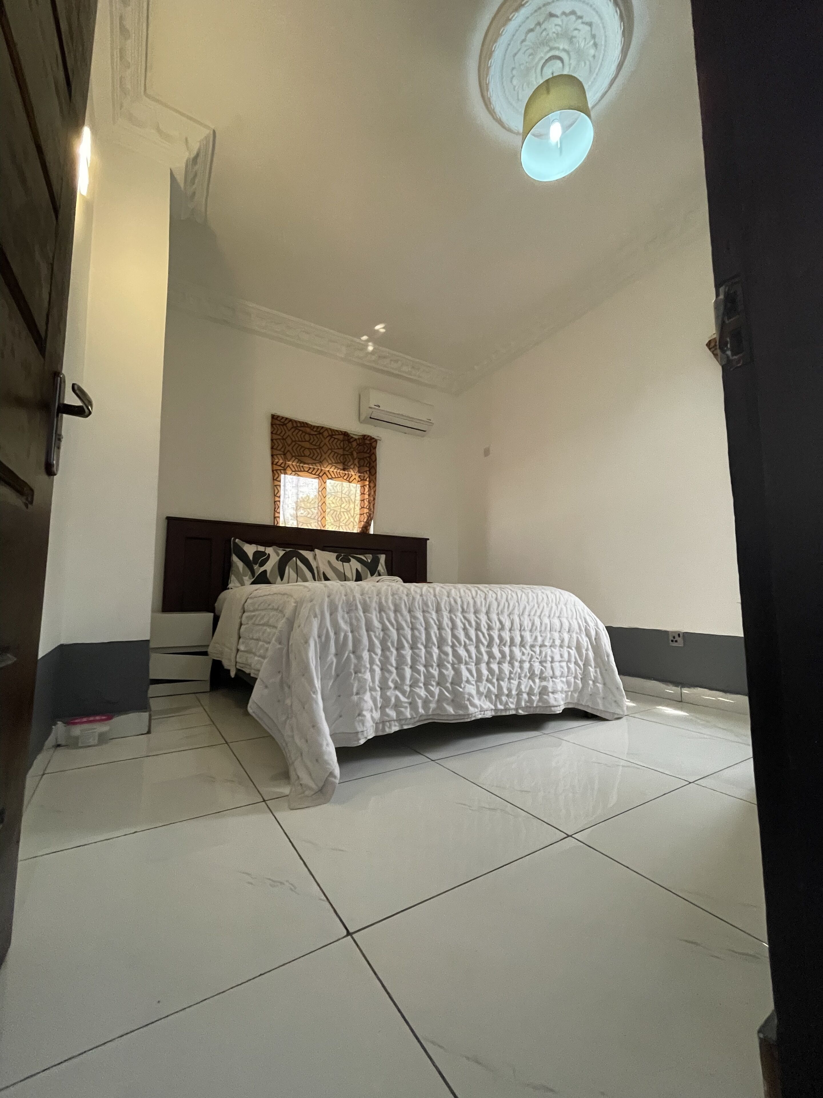Un dormitorio con cama, una ventana con cortinas estampadas, un equipo de aire acondicionado y un accesorio de iluminación en el techo.