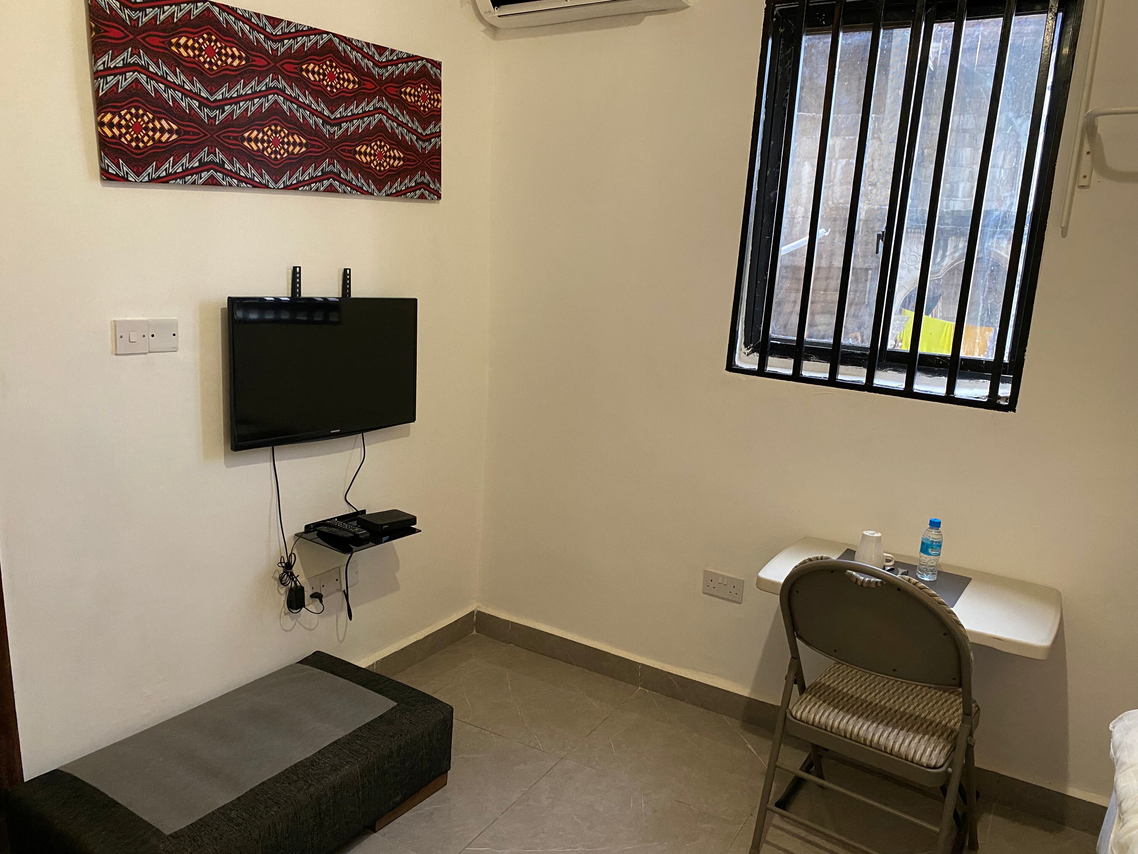Una habitación pequeña y bien iluminada, con una televisión de pantalla plana montada en la pared, una ventana con rejas, un escritorio pequeño con una silla y un banco.