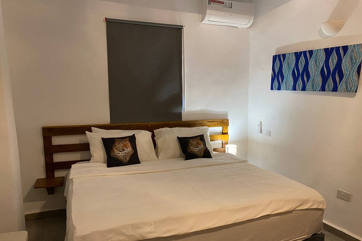 Een hotelkamer met een bed, hoofdbord, raam, airconditioning en een wandlamp, plus een kamerkunstwerk met een patroon.