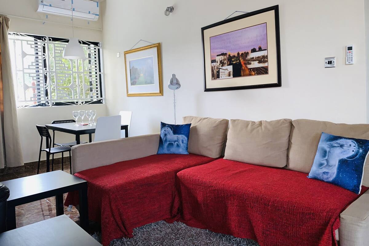 Een woonkamer met een beige bank, rode plaids en blauwe kussens. Er hangt een lijst met een afbeelding aan de muur, een tafel met stoelen en een raam met jaloezieën.