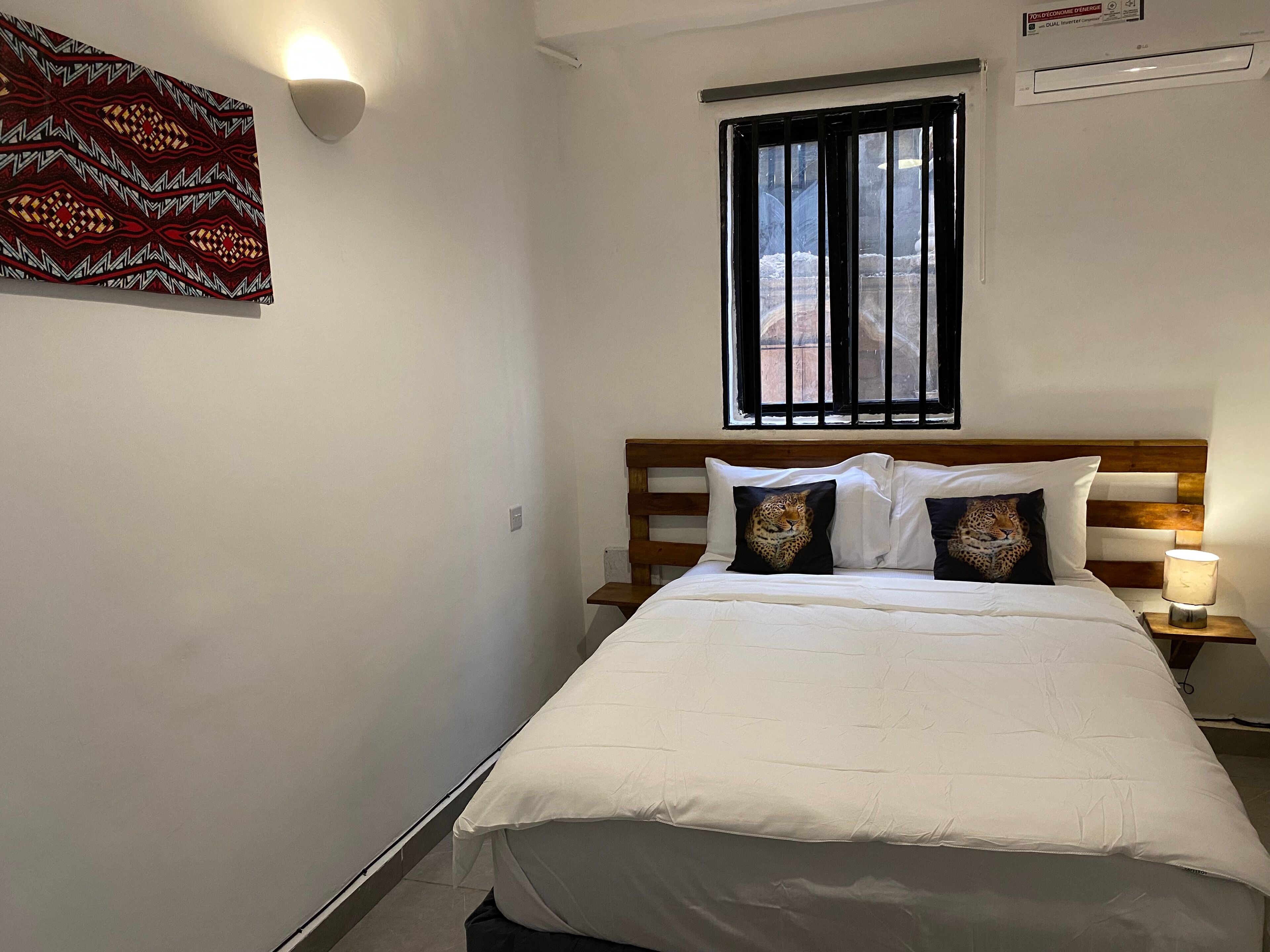 A bedroom with a bed, a nightstand with a lamp, a window with black bars, and a wall with a patterned artwork.