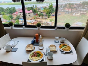 Una mesa de desayuno con platos de comida, una botella y tazas, frente a una ventana que ofrece vistas de un paisaje urbano.