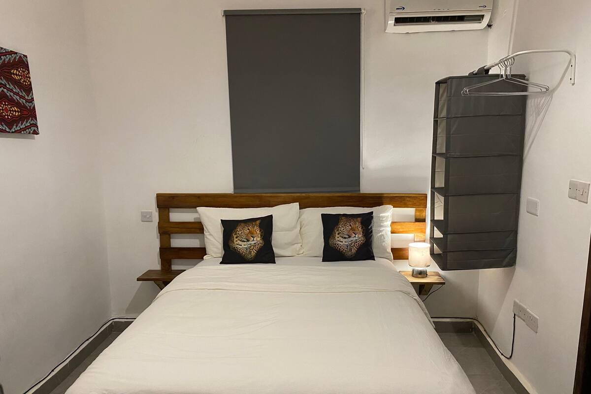 Een kleine, smalle hotelkamer met een houten hoofdbord, twee kussens met luipaardprint, een witte dekbedovertrek, een bedlamp en een wandairconditioning.
