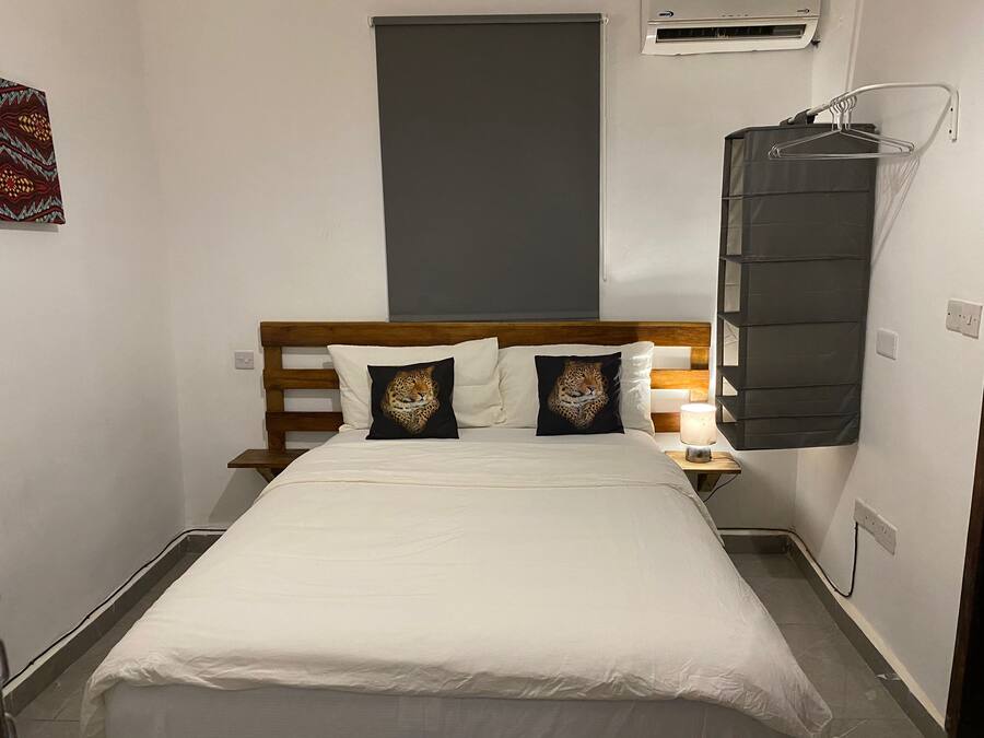 A small, narrow hotel room with a wooden headboard, two leopard-themed pillows, a white bedspread, a bedside lamp, and a wall-mounted air conditioning unit.