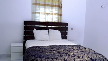 Un dormitorio con cama, mesitas de noche blancas, cabecero de madera y una ventana con cortinas.