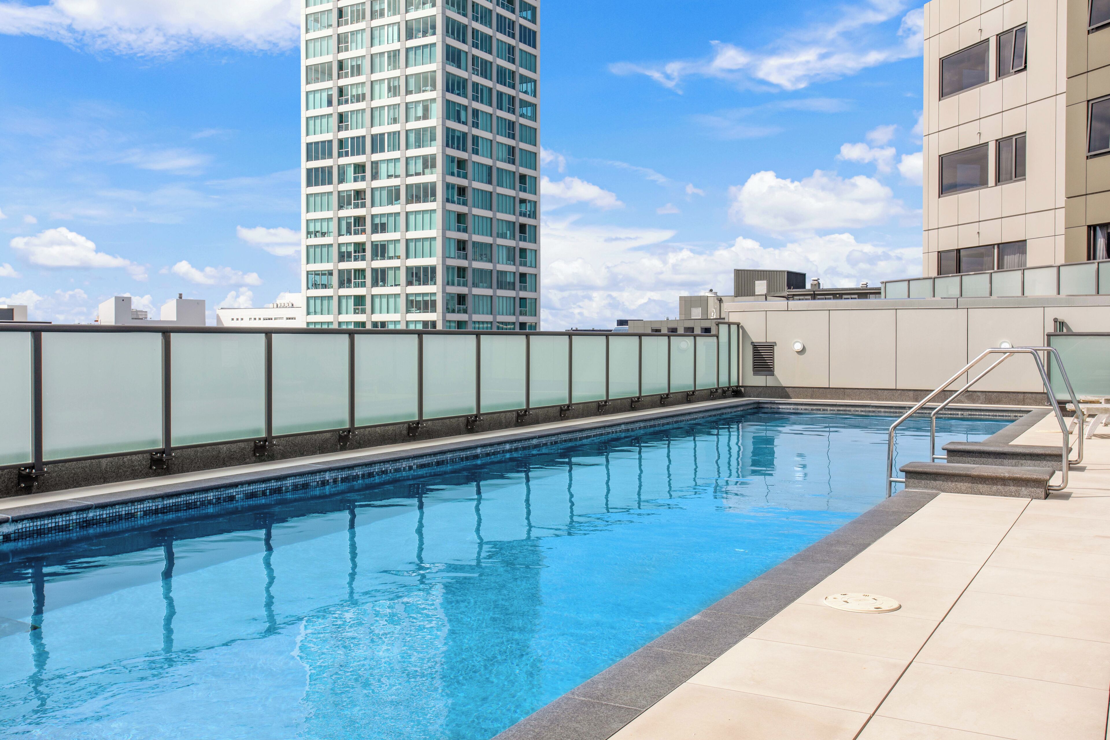 Una piscina en la azotea con barandilla de cristal, rodeada de edificios modernos y un cielo despejado.