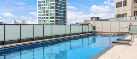 Una piscina en la azotea con barandilla de cristal, rodeada de edificios modernos y un cielo despejado.