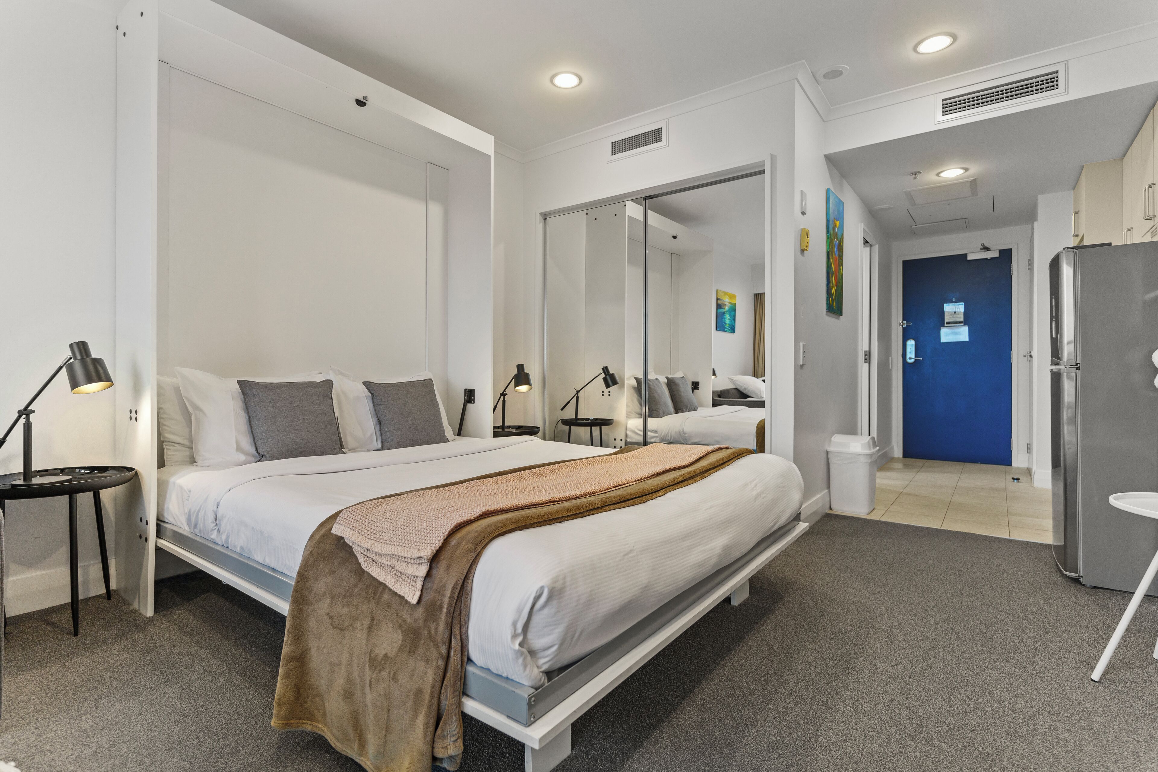 Una habitación de hotel moderna con una cama grande, mesitas de noche, un refrigerador y un baño visible a través de una puerta de cristal.