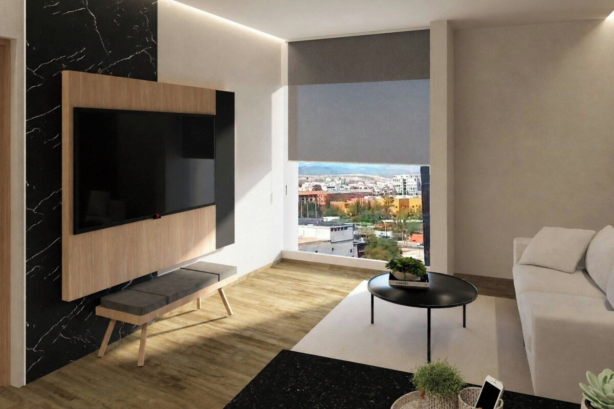 Una sala moderna con un televisor de pantalla plana montado en una pared de madera, una mesa de centro negra con una planta, un sofá blanco y un amplio ventanal con vistas a la ciudad.