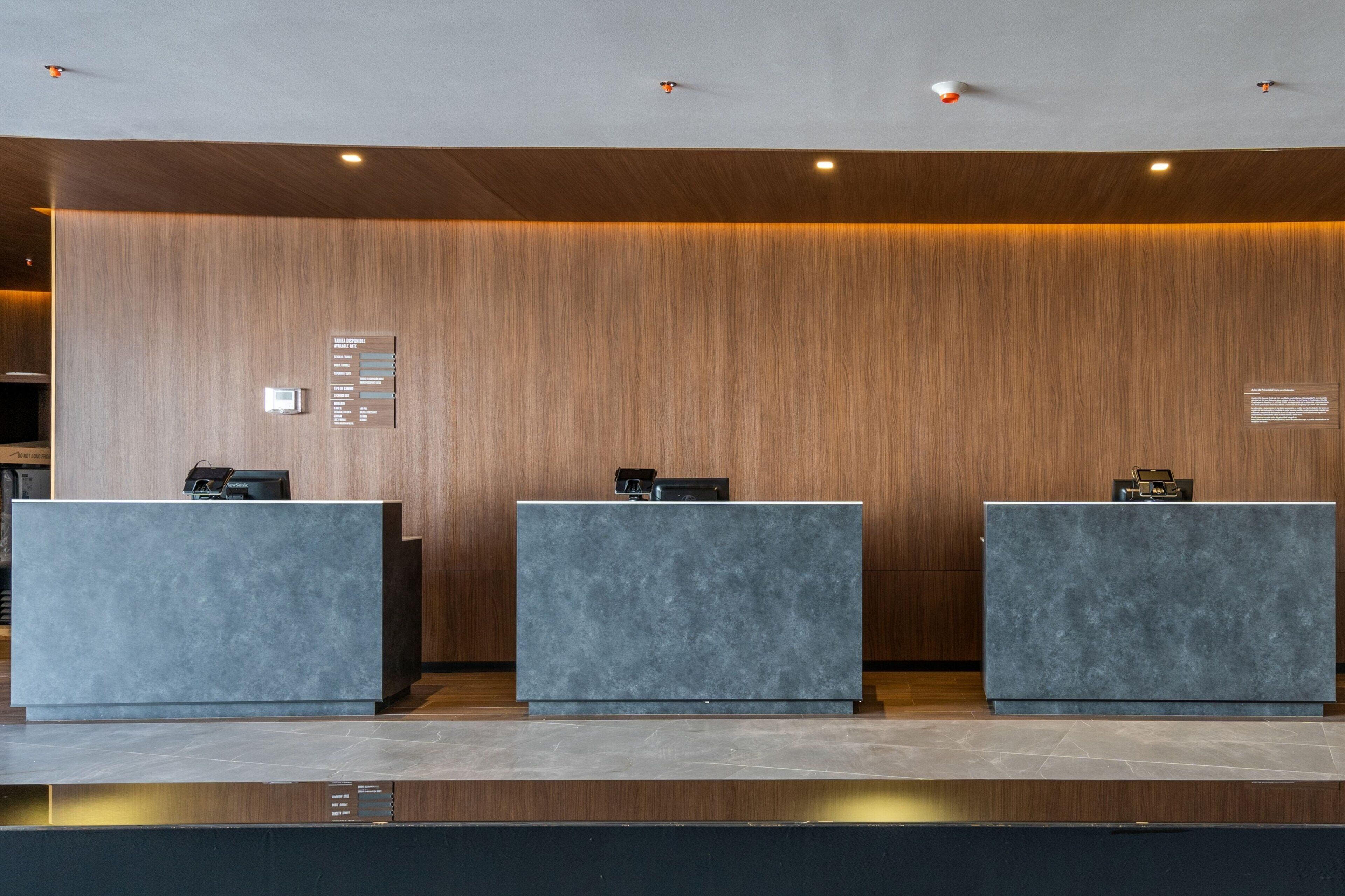A reception area with three grey reception desks against a wooden wall, equipped with a digital information board and a small clock.