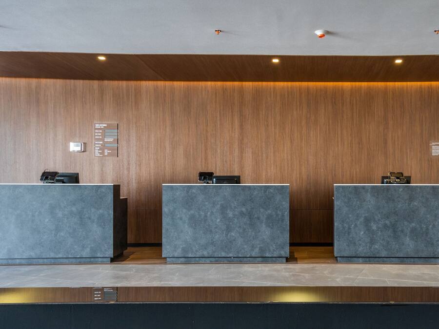 A reception area with three grey reception desks against a wooden wall, equipped with a digital information board and a small clock.