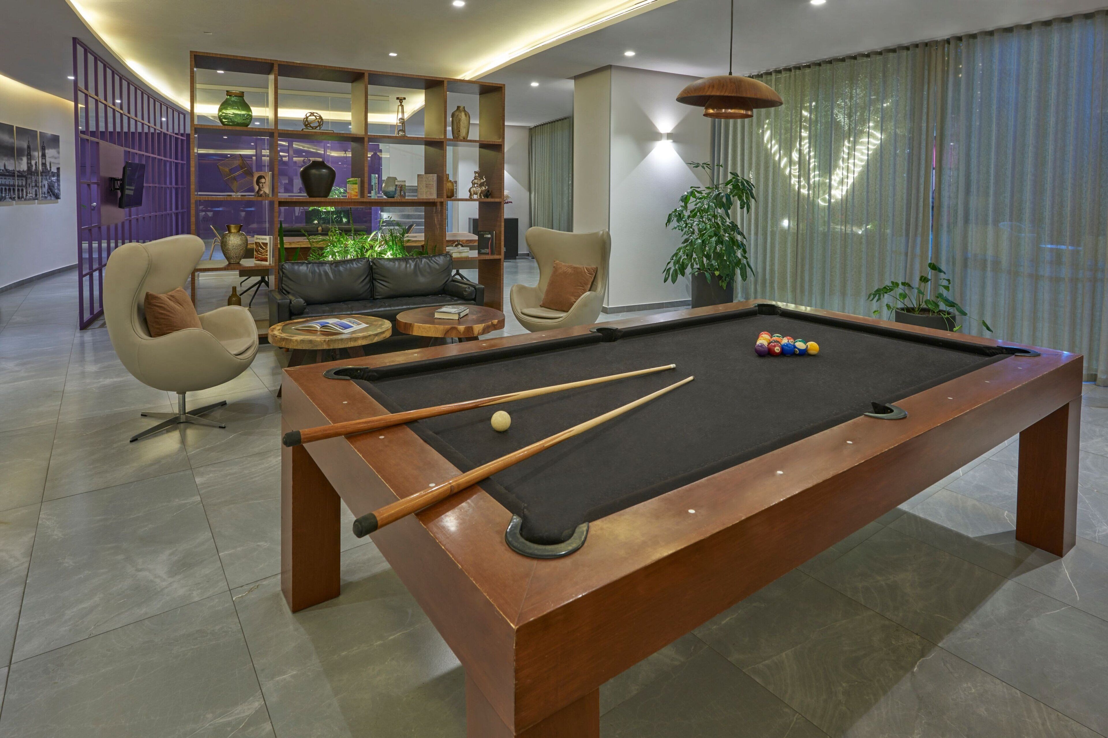 A pool table with billiard balls, a sofa, and a coffee table in a modern living room with a bookshelf and a potted plant.