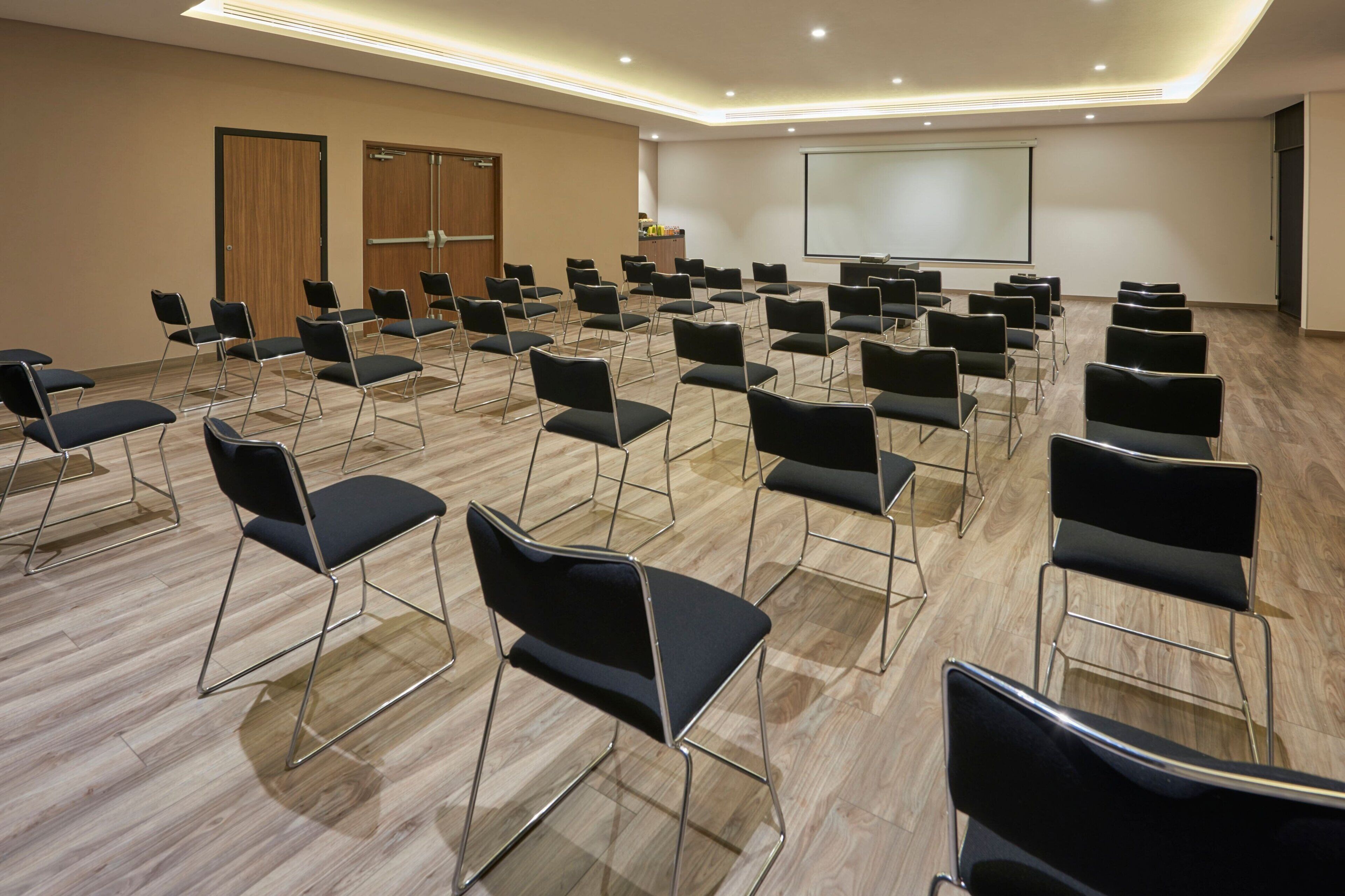 Un salón de conferencias con filas de sillas negras, una pantalla de proyector y un atril.