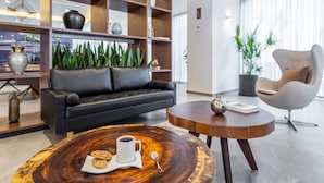 Una sala de estar moderna con un sofá de cuero negro, una mesa de centro redonda de madera con una taza y galletas, un sillón blanco y una estantería llena de objetos decorativos.