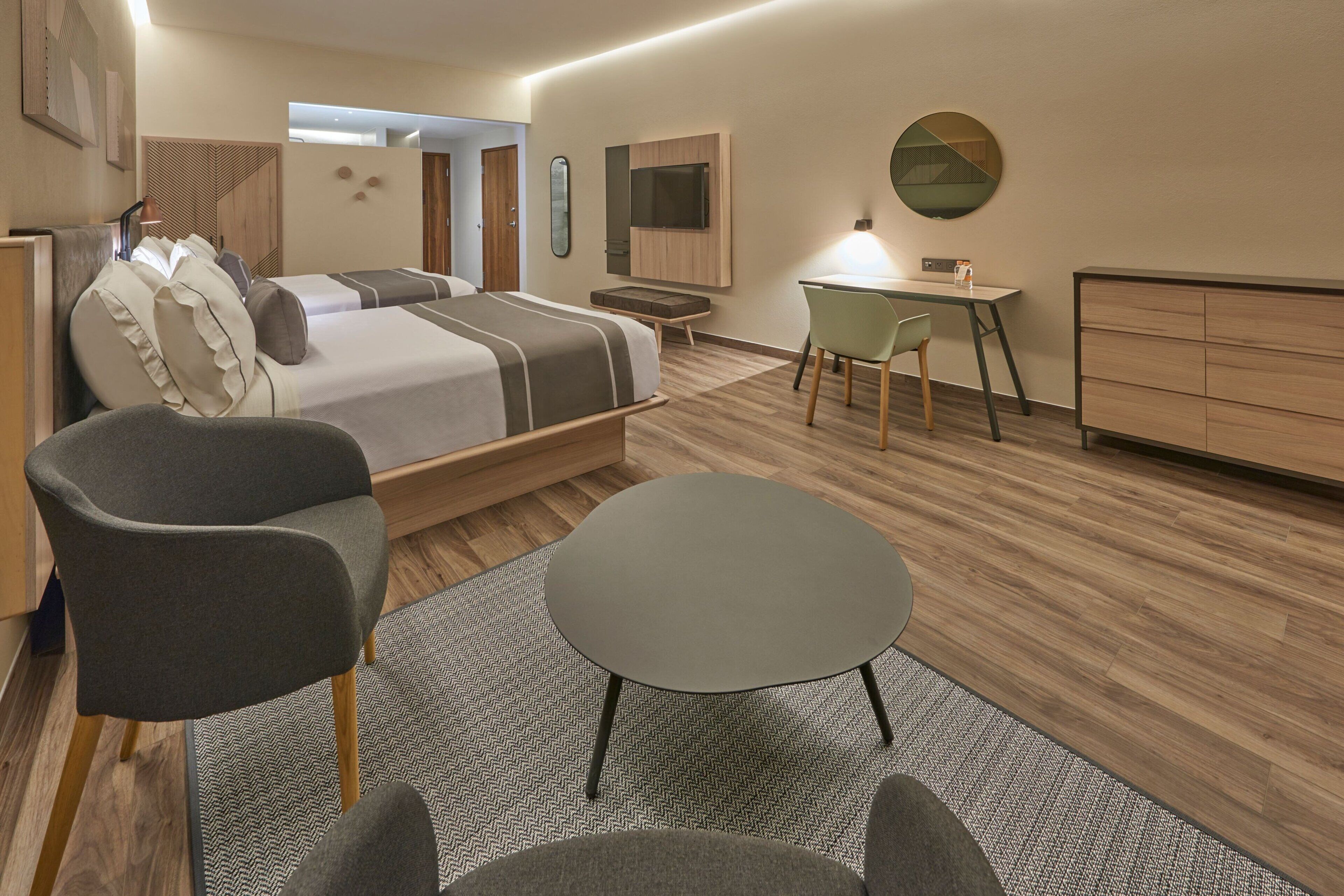 A modern hotel room with a bed, two armchairs, a desk, and a dresser.