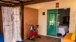 A room with a wooden floor, a red stationary bike, a sliding glass door leading to a balcony, and a bed with a white sheet.