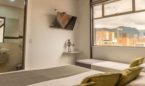 Habitación de hotel moderna con cama, un pequeño estante con una escultura, televisor montado en la pared y un gran ventanal con vistas a la ciudad.