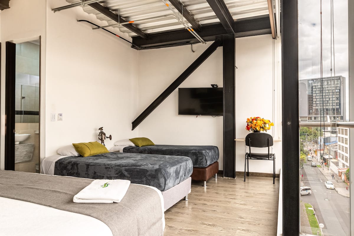 Una habitación de hotel moderna con dos camas, televisor, ventana con vista a la ciudad y balcón con una silla y flores.