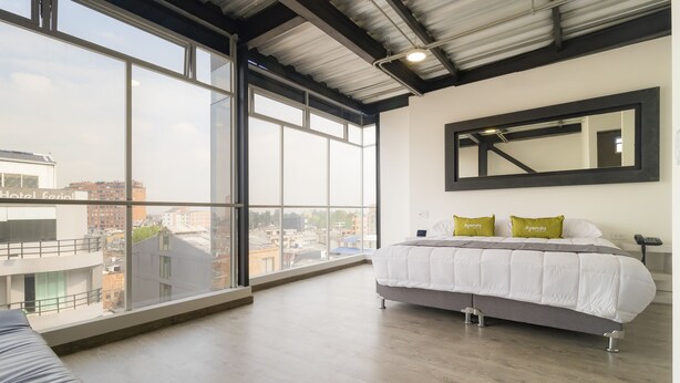 A hotel room with a large window, a bed with white bedding and two pillows, a black-framed mirror, and a wooden floor. - Hotel Wess Bogota (Bogotá)