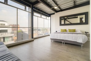 Habitación de hotel con un ventanal grande, cama con ropa blanca y dos almohadas, espejo con marco negro y piso de madera.