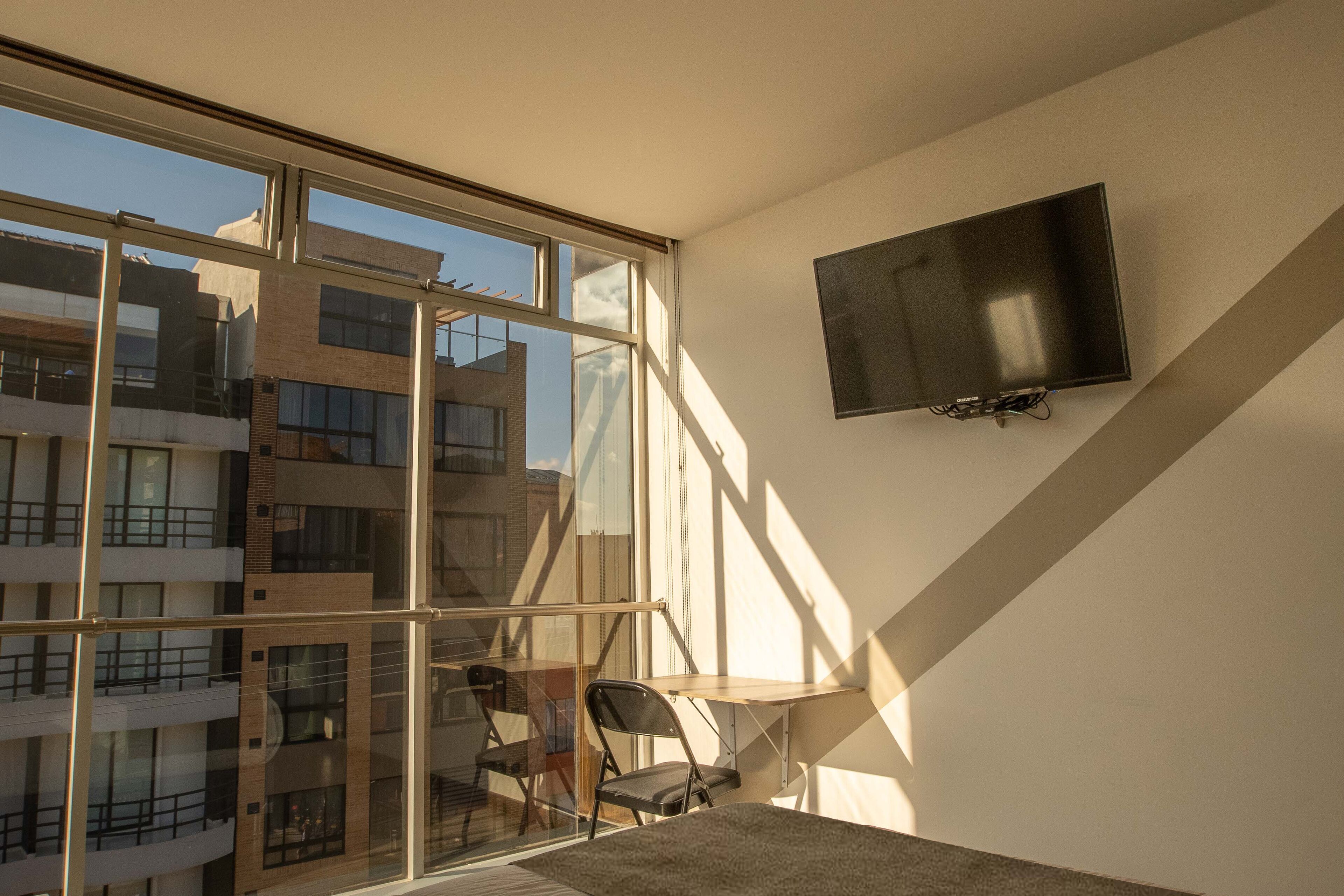 Une chambre moderne dotée d’une grande fenêtre, d’un téléviseur à écran plat fixé au mur et d’une petite table avec des chaises.