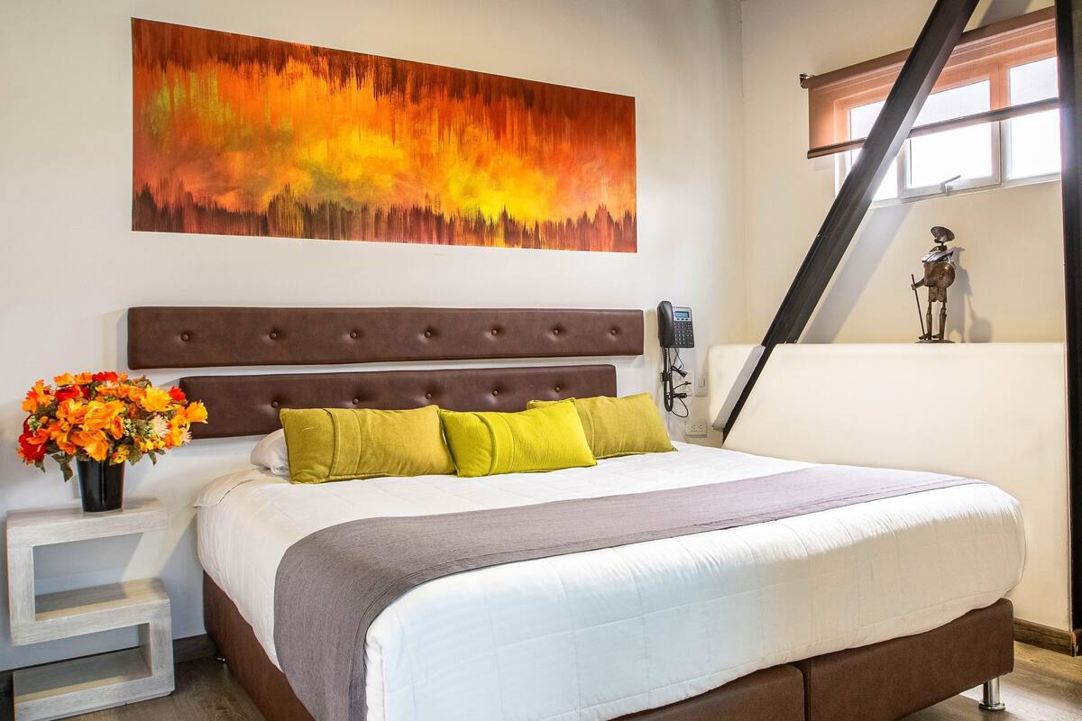 Un dormitorio con una cama grande, un cabecero, una mesita de noche con un florero y un cuadro en la pared.