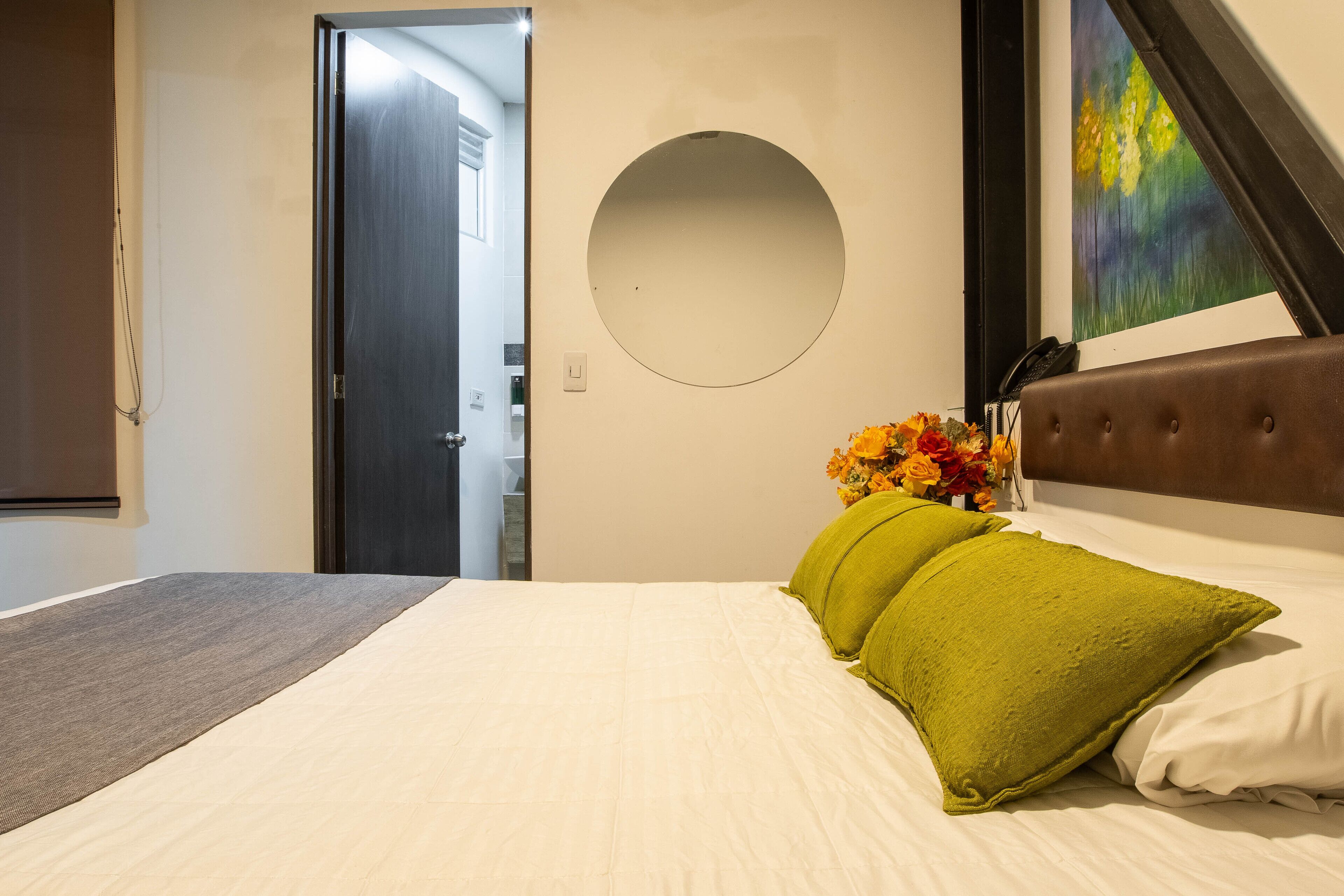 A hotel room with a bed, a round mirror, a painting, and a vase with flowers.
