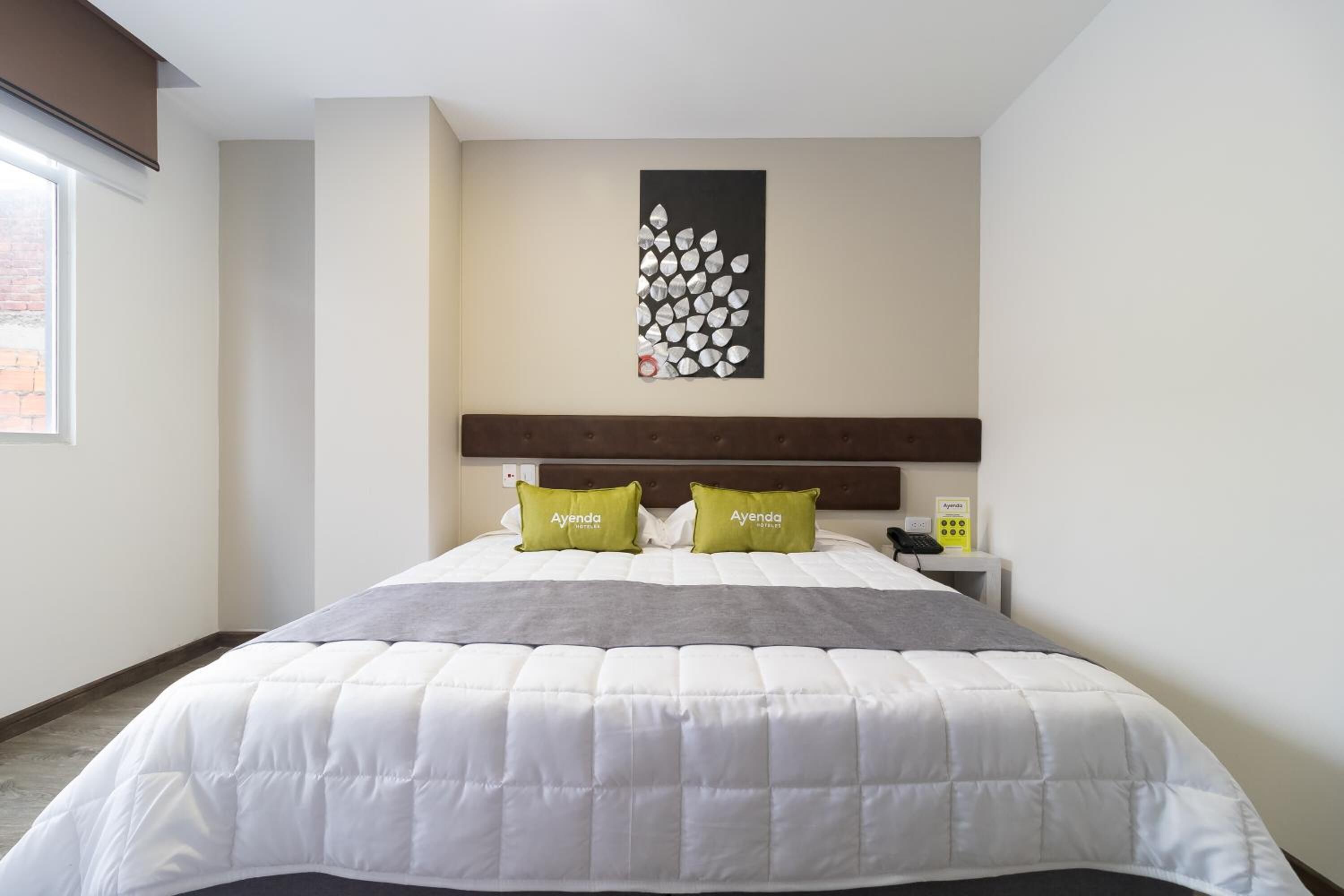 A hotel room with a large bed, two green pillows, a headboard, a bedside table with a phone and a small device, and a wall art piece.