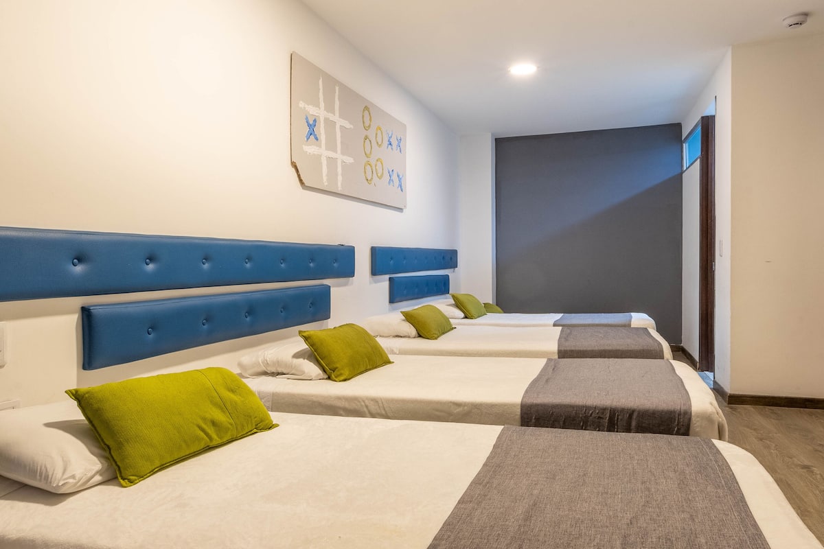 Habitación de hotel con cuatro camas, cada una con un cabecero de color diferente y una almohada verde.