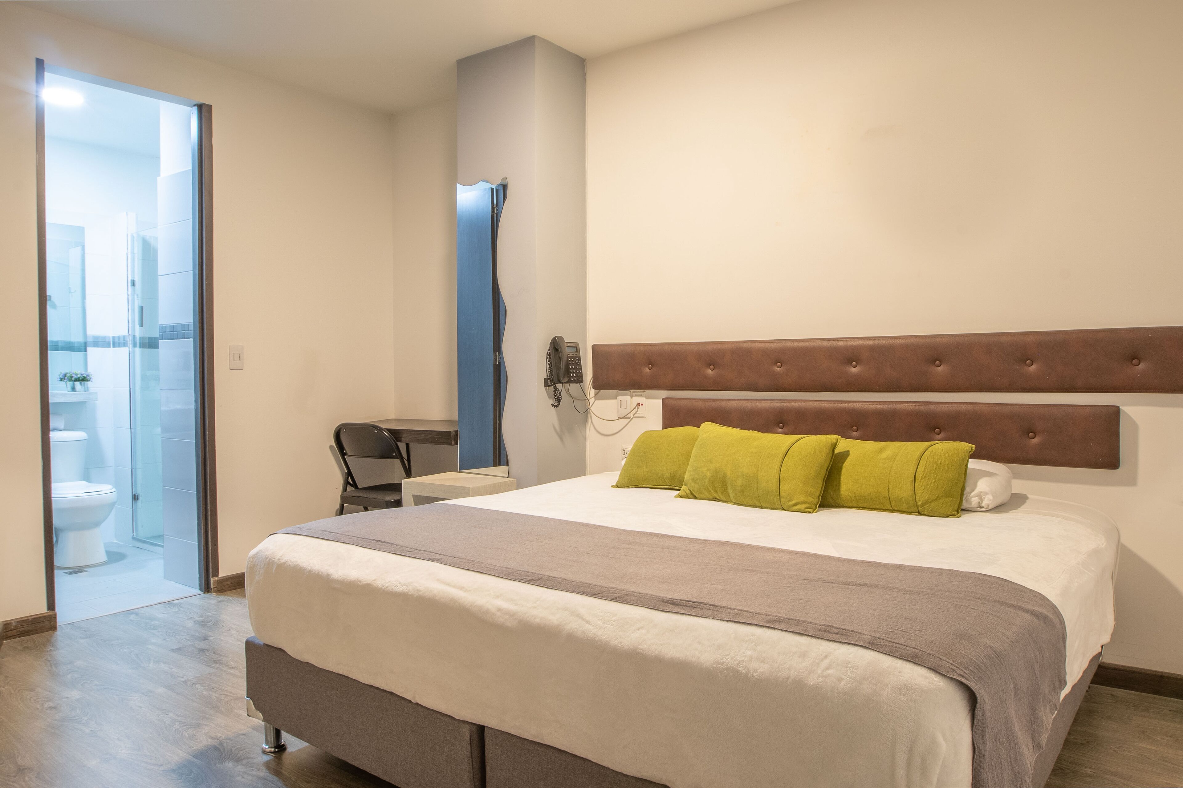 A hotel room with a bed, a bedside table, a chair, and a bathroom visible through a glass door.
