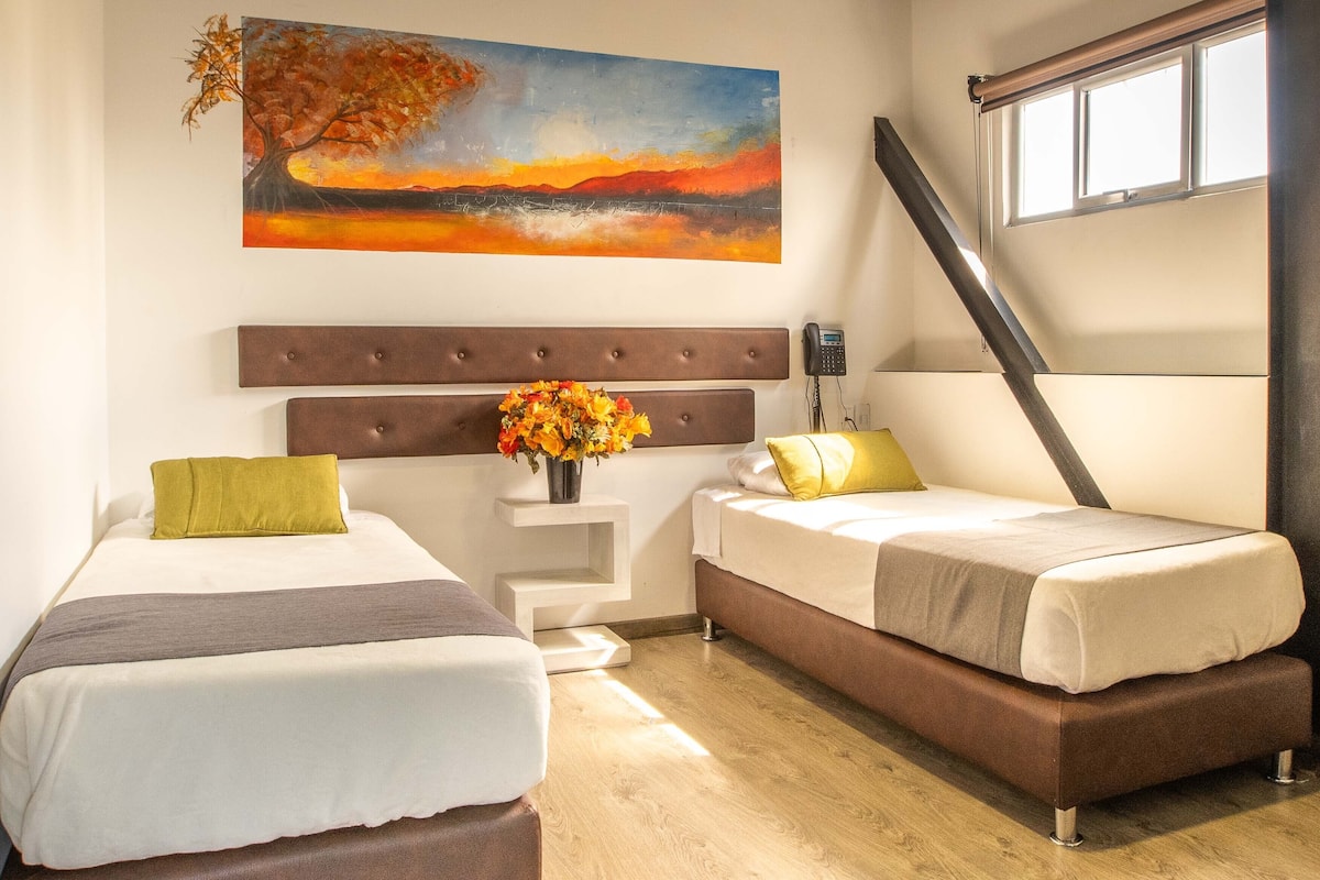 Una habitación de hotel con dos camas, un cuadro en la pared, un florero con flores y una ventana con persianas.