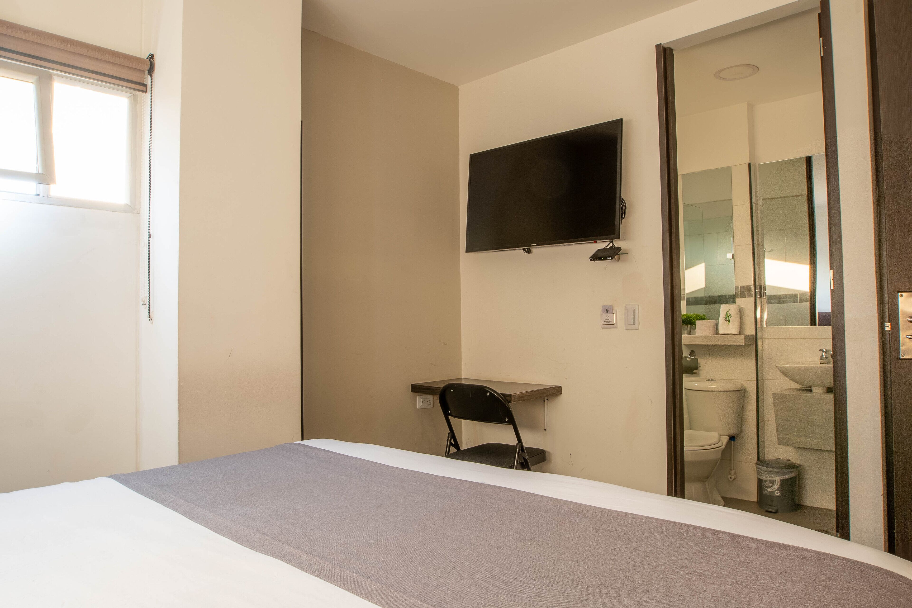 A hotel room with a bed, a TV mounted on the wall, a chair, and a bathroom visible through an open door.