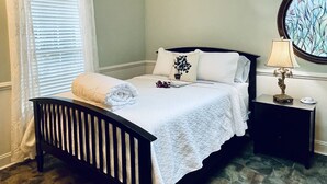 A bedroom with a skylight, a bed with white linens, a nightstand with a lamp, and a window with blinds.