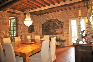 A stone-walled dining area with a wooden table and white chairs, a chandelier, and a rustic fireplace.