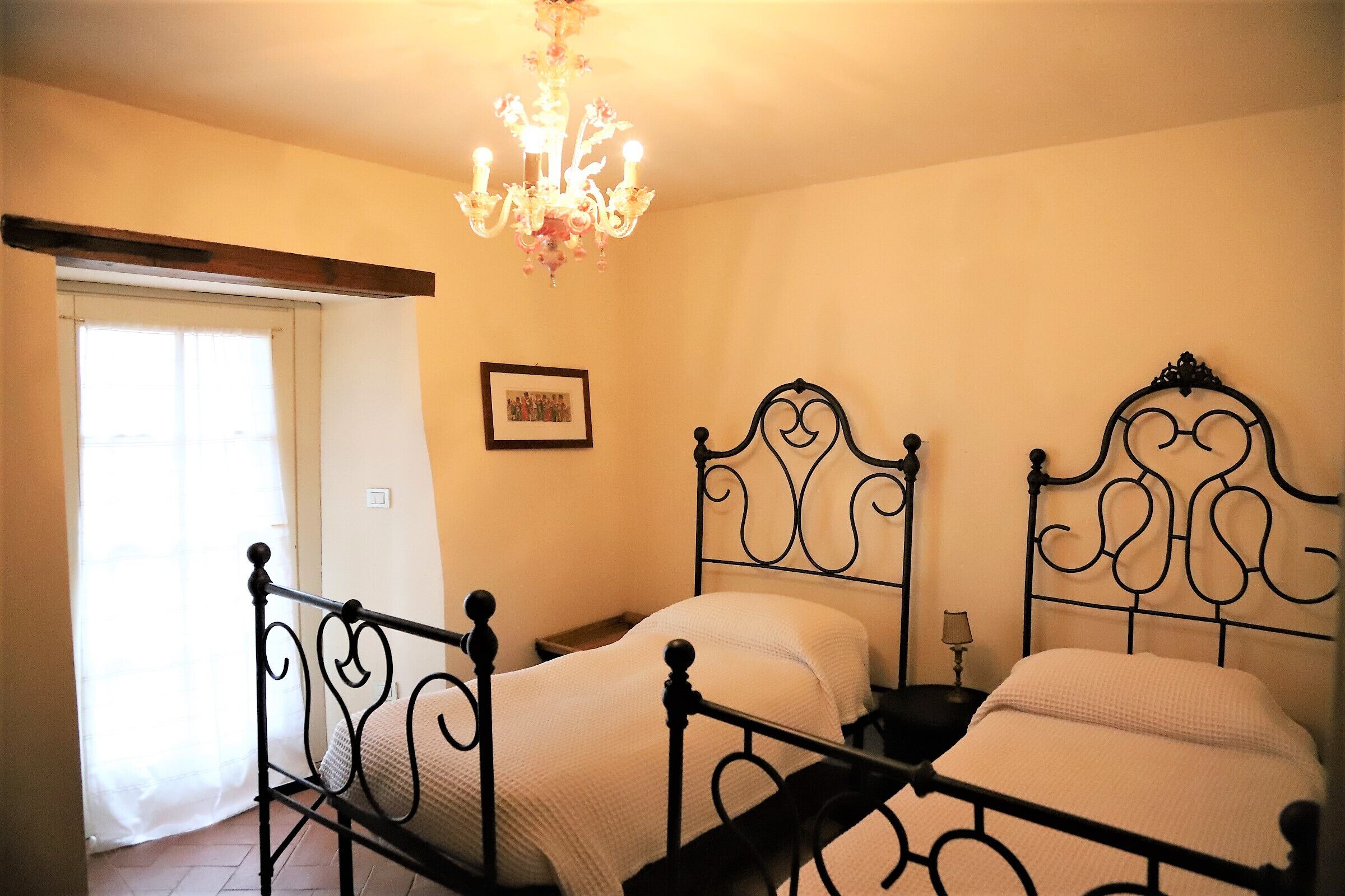 A bedroom with two beds, a chandelier, a picture on the wall, and a window with curtains.
