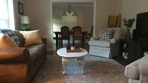 Una sala de estar con un sofá de cuero marrón, una mesa de centro blanca y un sillón beige. Se observa un comedor al fondo con una araña.