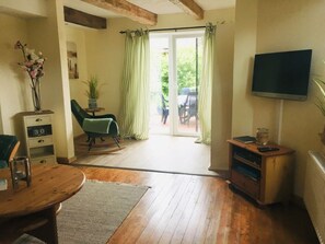 Un salon chaleureux avec un plancher en bois, un fauteuil vert, une petite table, un téléviseur et une porte-fenêtre coulissante donnant sur un espace extérieur.