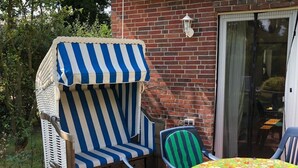 A patio with a striped beach chair, a table with a floral tablecloth, and a brick building with a striped awning. - Restored Gulfhof in the heart of East Frisia (Ihlow)