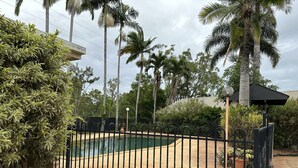 Una piscina rodeada por una cerca de metal negro, con palmeras y diversas plantas al fondo.